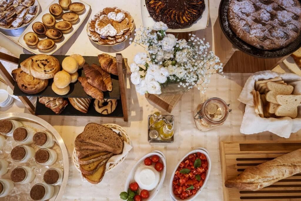 A variety of baked goods including pastries, bread, and a cake on a table with flowers and condiments.