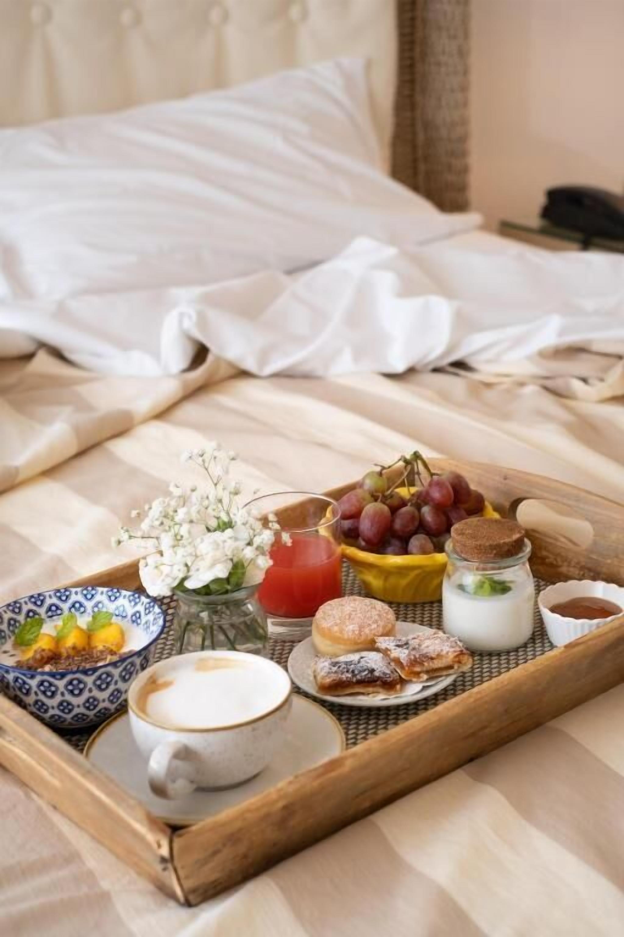 A breakfast tray on a bed with a cup of coffee, a glass of red juice, a bowl of grapes, a jar of milk, a small bowl of jam, and a plate of pastries.