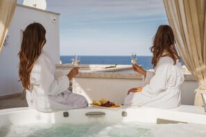 Two people in white robes sitting in a hot tub with a view of the sea, holding glasses of white wine, and a plate of fruit. - Relais Valle dell'Idro (Otranto)