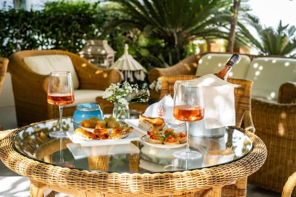 Una mesa de vidrio con comida y bebidas, mobiliario de mimbre y un entorno tropical al aire libre.
