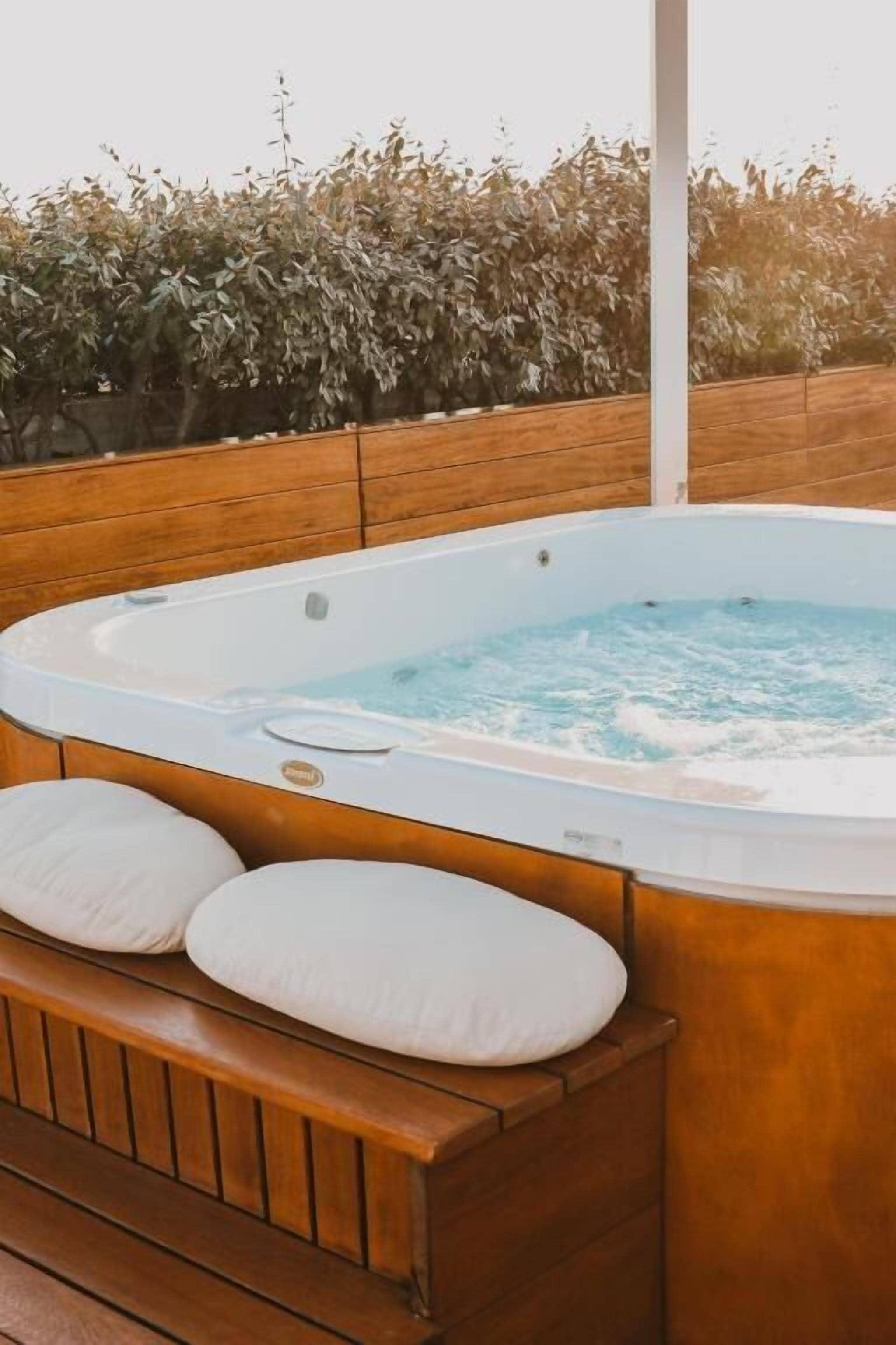 A hot tub with two white cushions, set against a wooden deck with a hedge in the background.