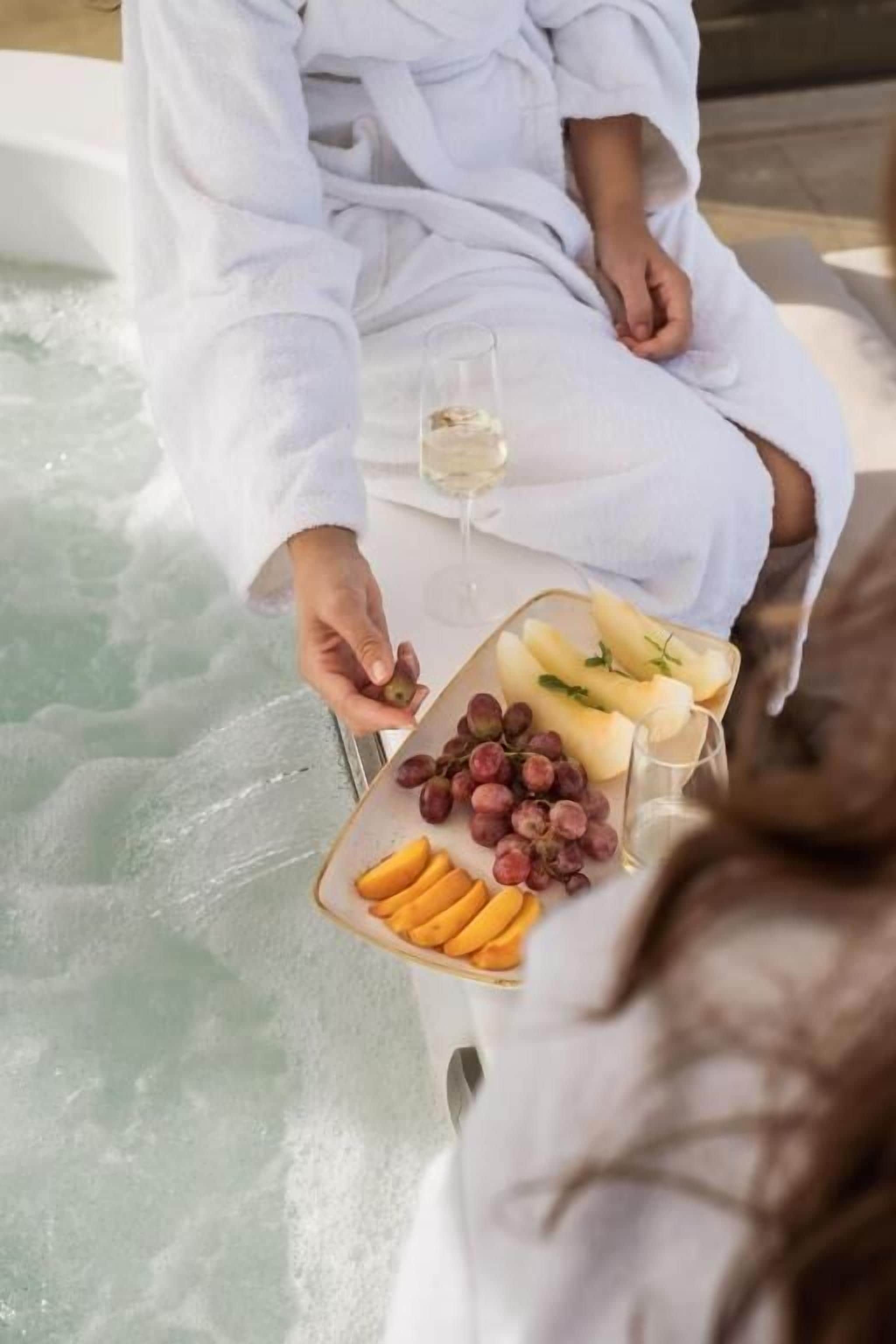 A person in a white bathrobe sitting by a hot tub with a tray of fruits and a glass of white wine.