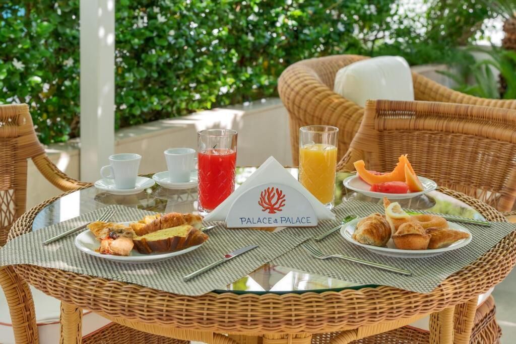 Une table en osier, dressée pour le petit-déjeuner, avec des assiettes remplies de nourriture, des verres de jus de fruits et une pancarte indiquant « Palace & Palace ».