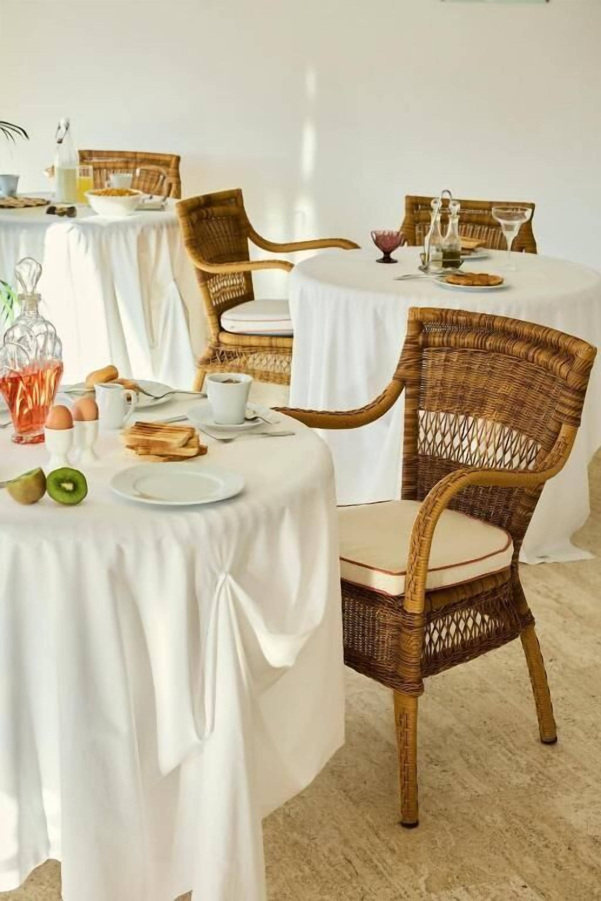 Un comedor con mesas redondas cubiertas con manteles blancos, sillas de mimbre y alimentos para el desayuno como huevos, tostadas y fruta.