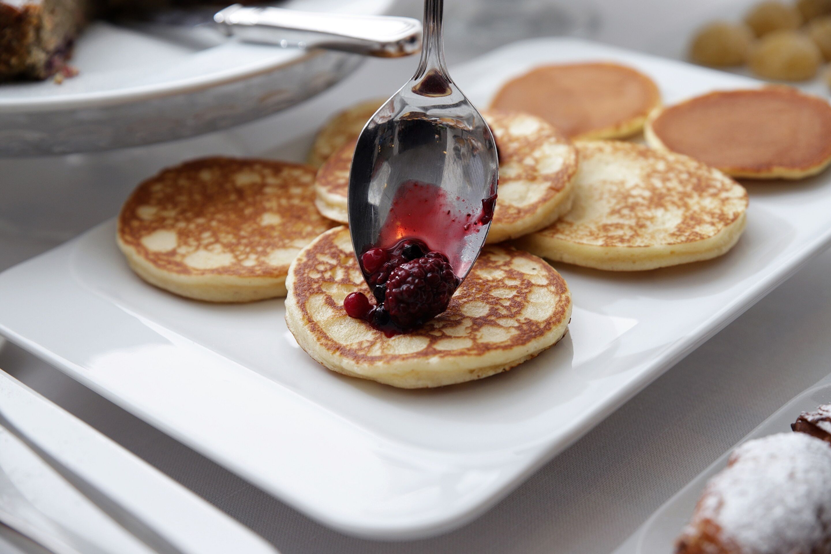 Une assiette rectangulaire blanche sur laquelle on voit plusieurs crêpes et une cuillère contenant une compote de fruits rouges.