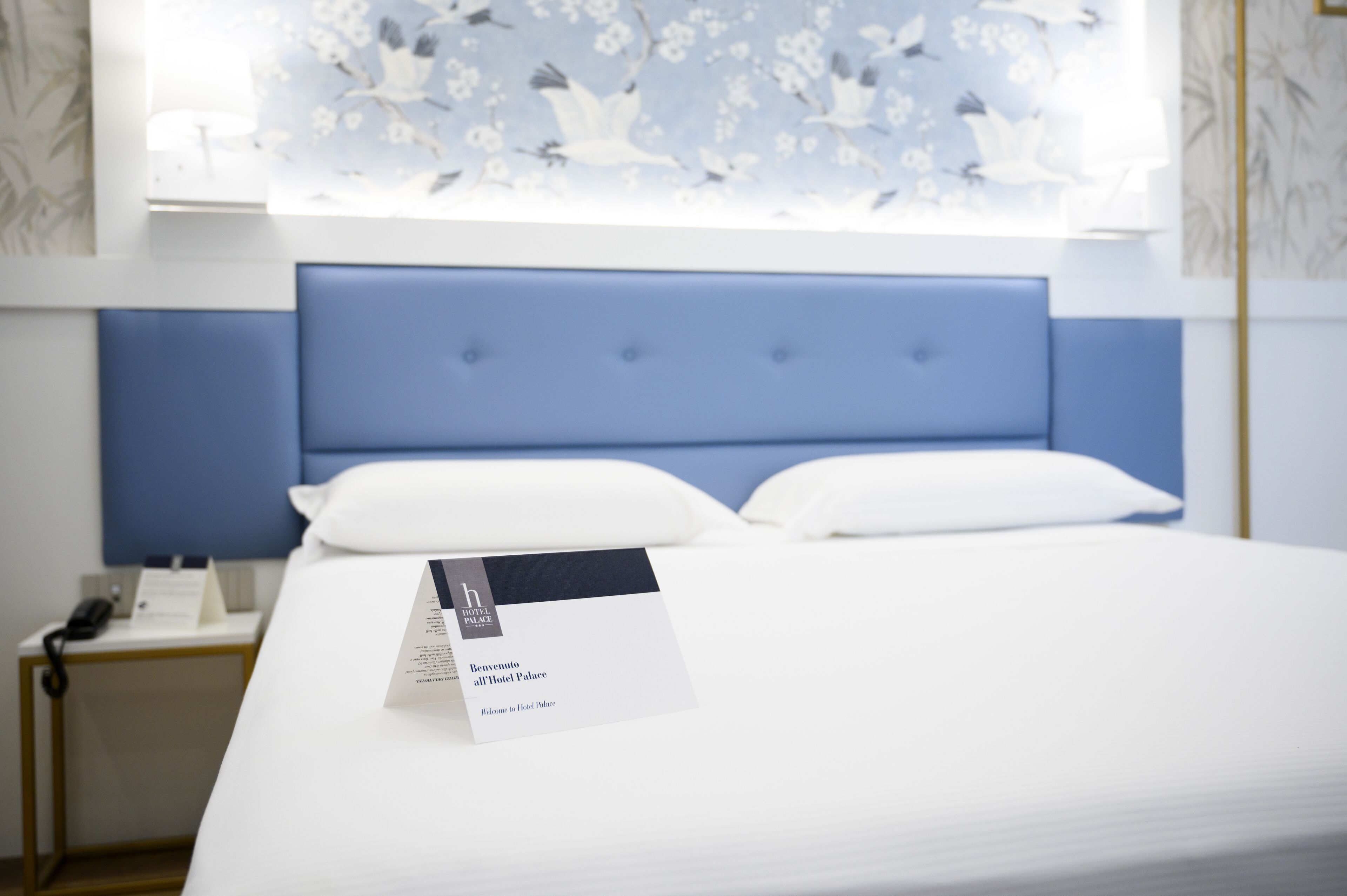 A hotel room with a bed, a bedside table with a phone, and a welcome card on the bed.