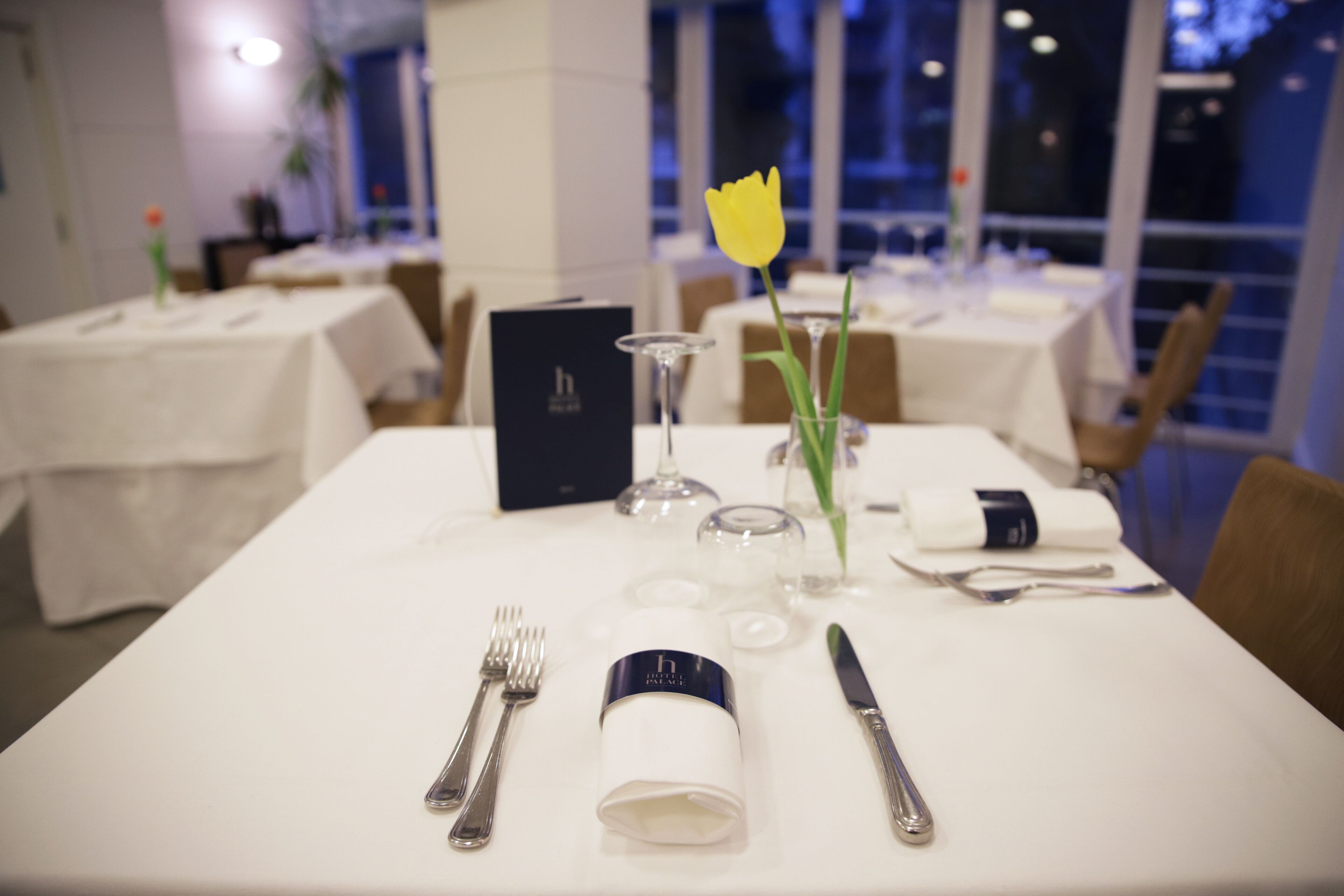 A dining table set with cutlery, a glass, a napkin, and a tulip in a vase.