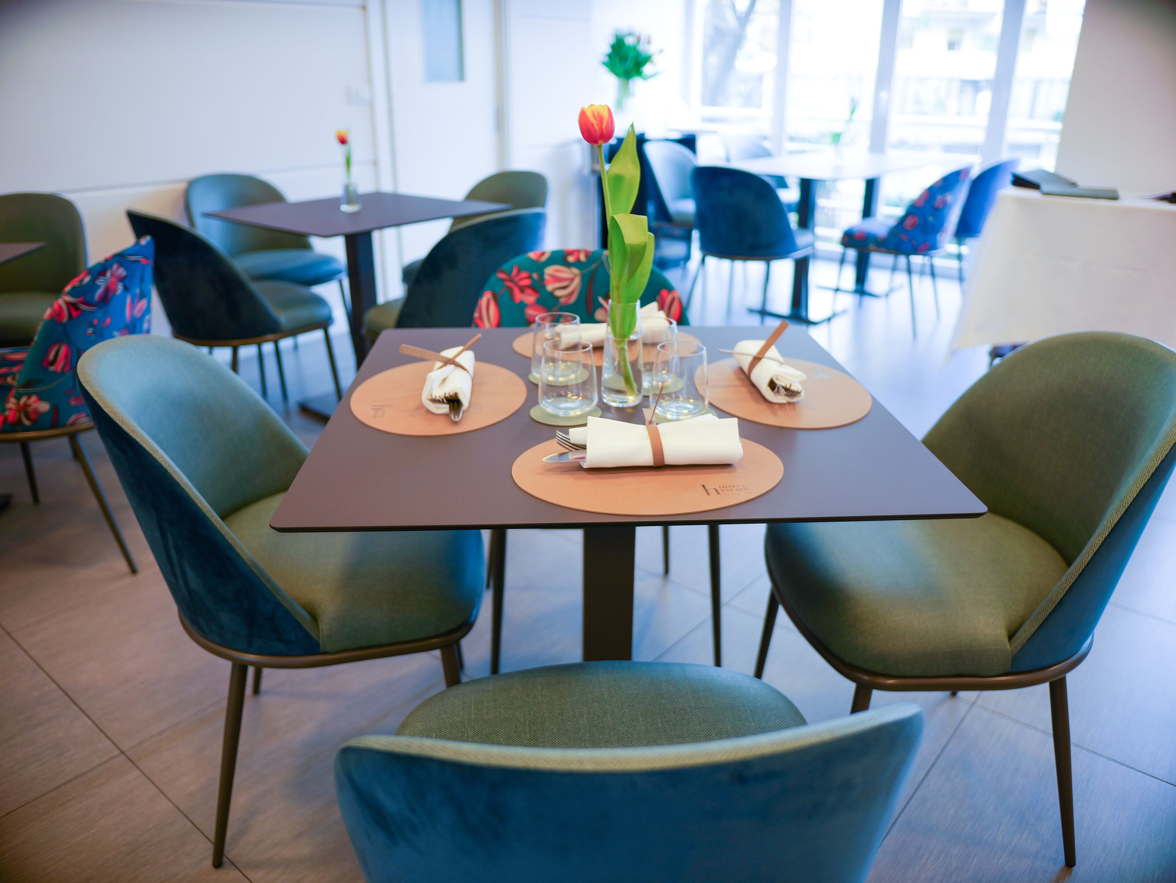 A dining area with a table set for two, featuring a vase with a single red tulip, two folded napkins, and a glass of water.
