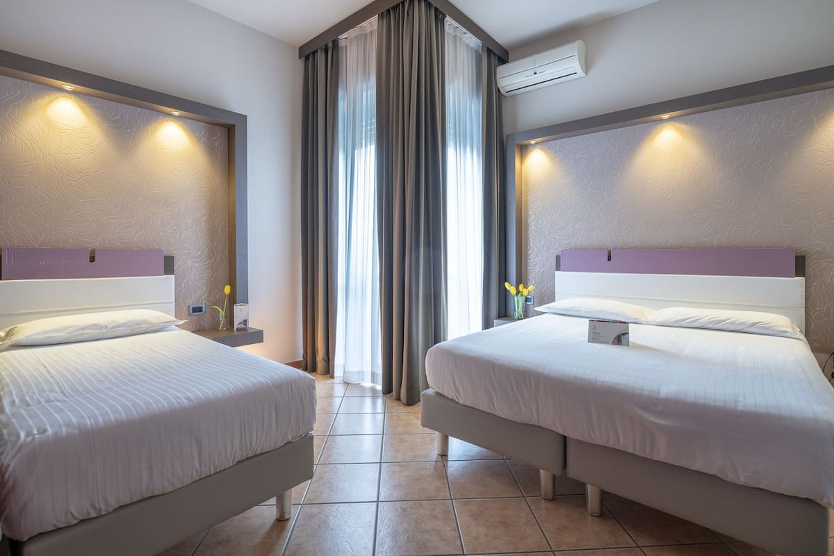 Ein Hotelzimmer mit zwei Betten, einem großen Fenster mit Vorhängen, einer Klimaanlage und einem kleinen Tisch mit Blumen in einer Vase.