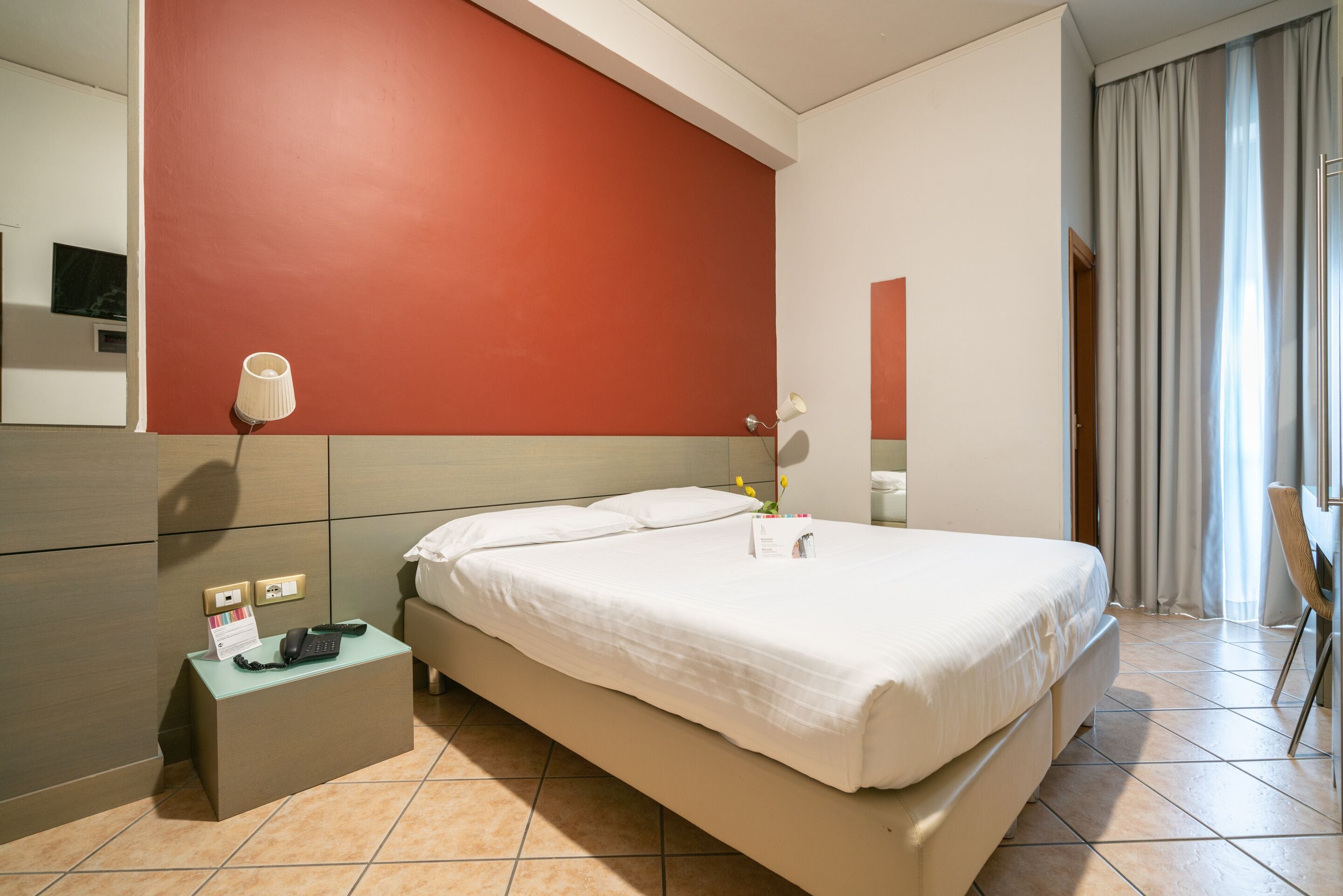A hotel room with a large bed, a bedside table with a lamp, a telephone, and a small table with a flower vase.