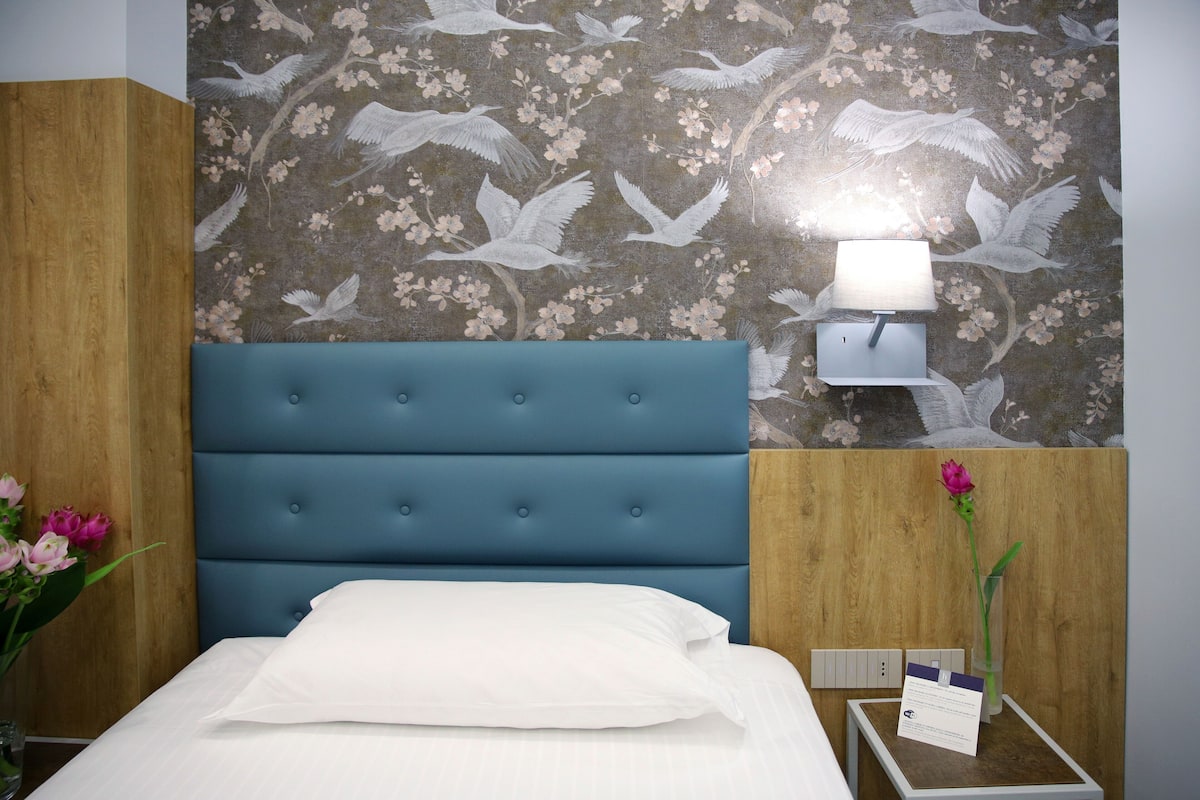 Ein Schlafzimmer mit blauem Kopfteil, einem weißen Bett, einer Wandlampe, einem Holznachttisch mit Blumenvase und floralem Wandpapier.