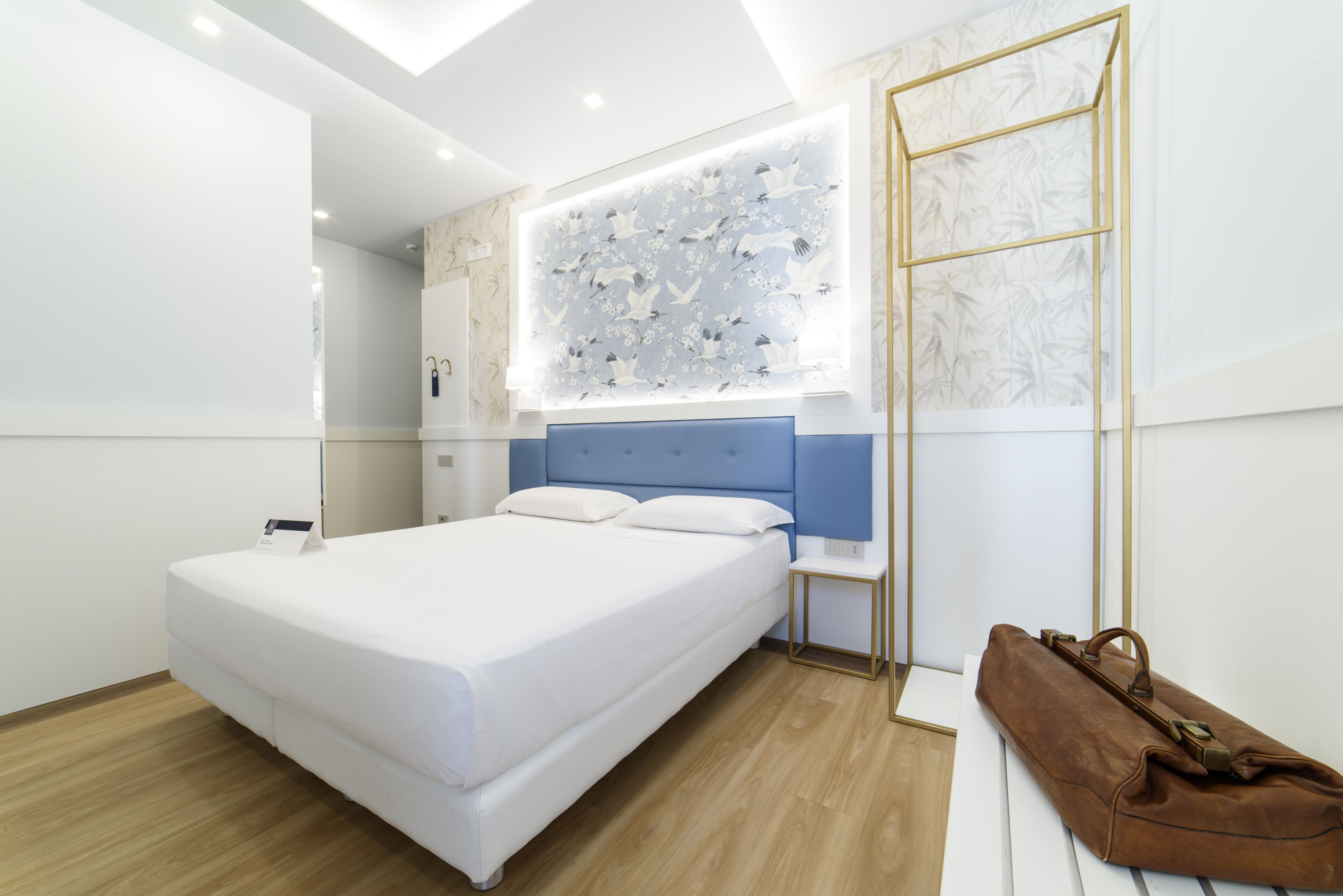 A modern hotel room with a large bed, a blue headboard, a wall-mounted artwork, a wooden floor, and a brown leather bag on a white bench.