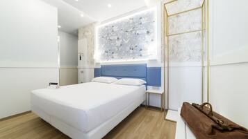 Una camera d'albergo moderna con un letto grande, una testiera blu, un'opera d'arte appesa al muro, un pavimento in legno e una borsa in pelle marrone su una panca bianca.