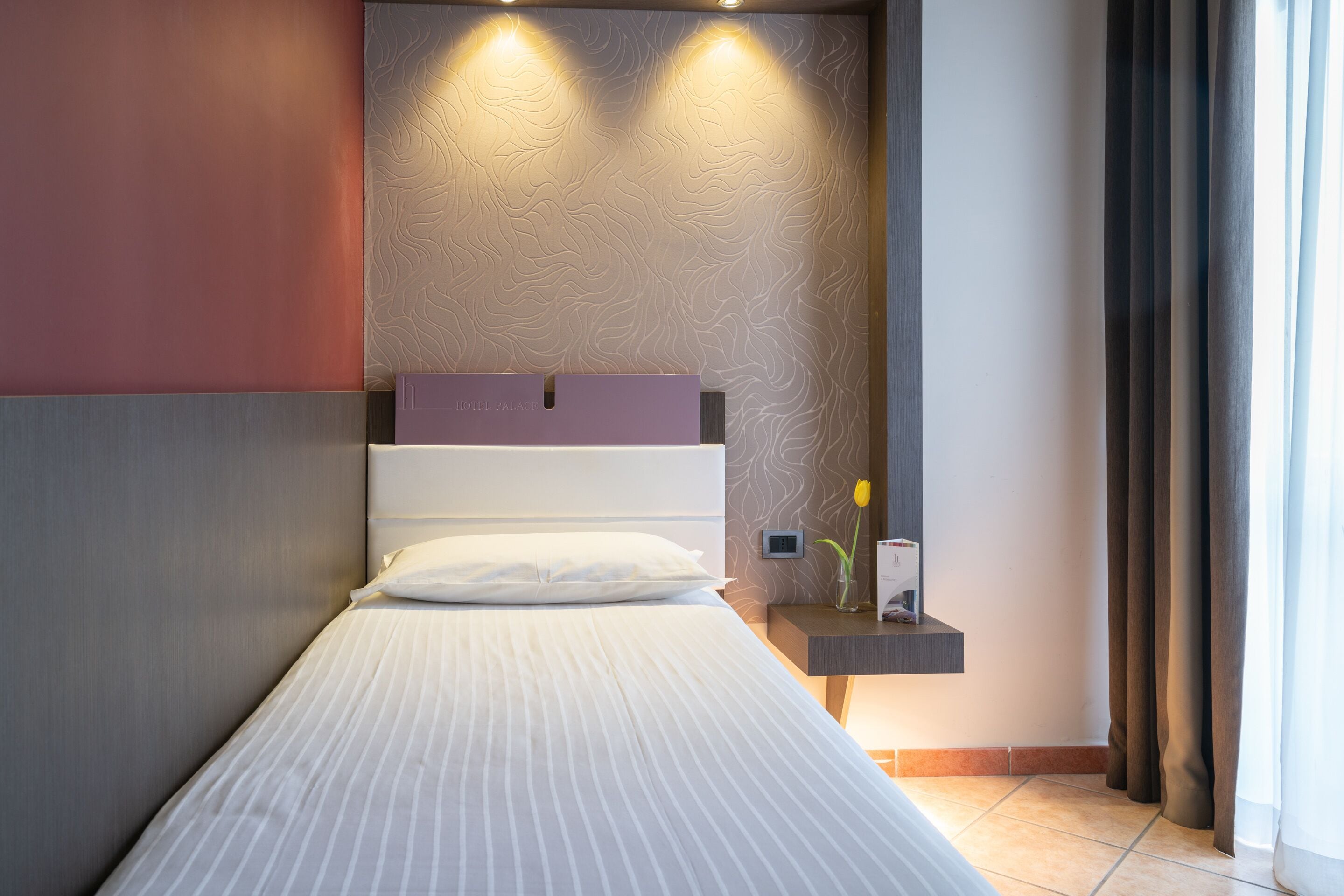 A neatly made bed with a headboard, a bedside table with a vase and a book, and a wall with a textured pattern.
