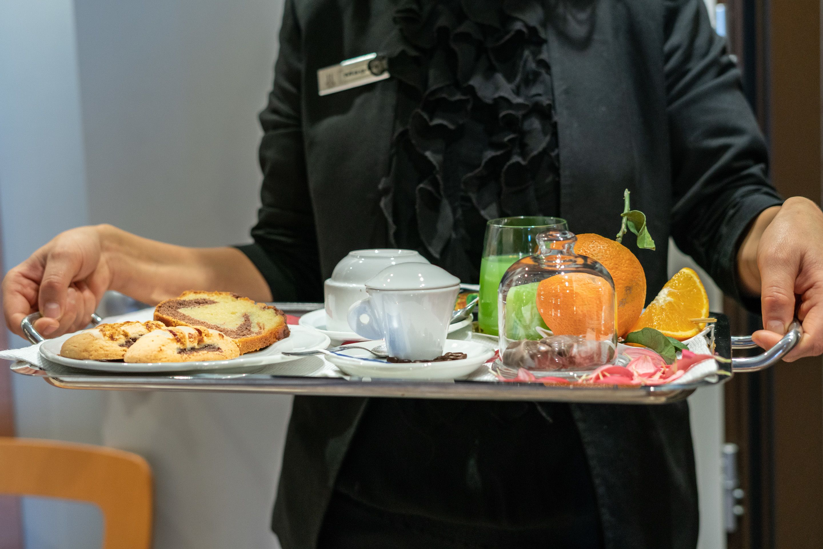 A person holding a tray with a teapot, a glass of green drink, an orange, and a slice of cake.