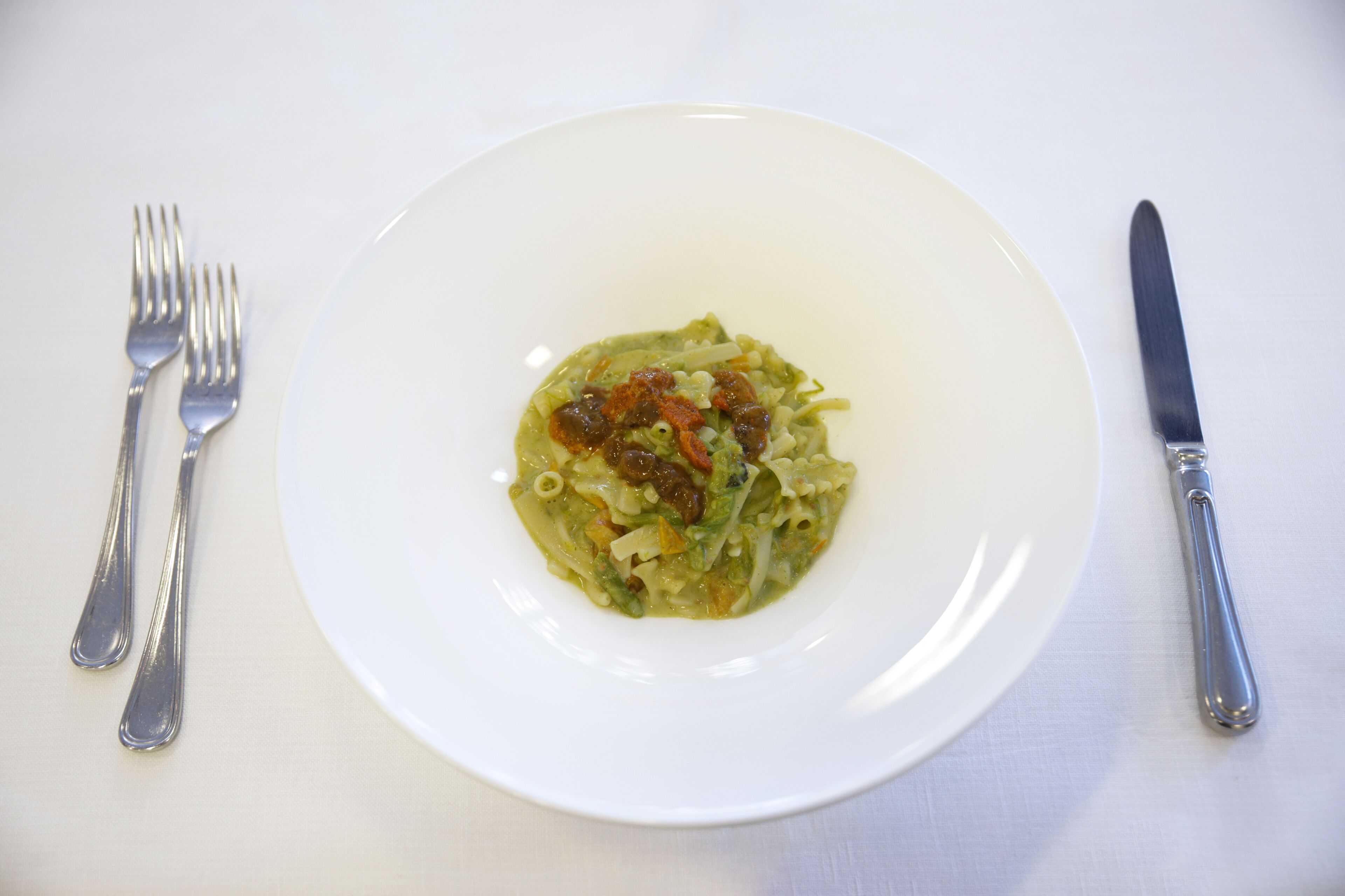A white plate with a green pasta dish, accompanied by a fork and knife on a white tablecloth.
