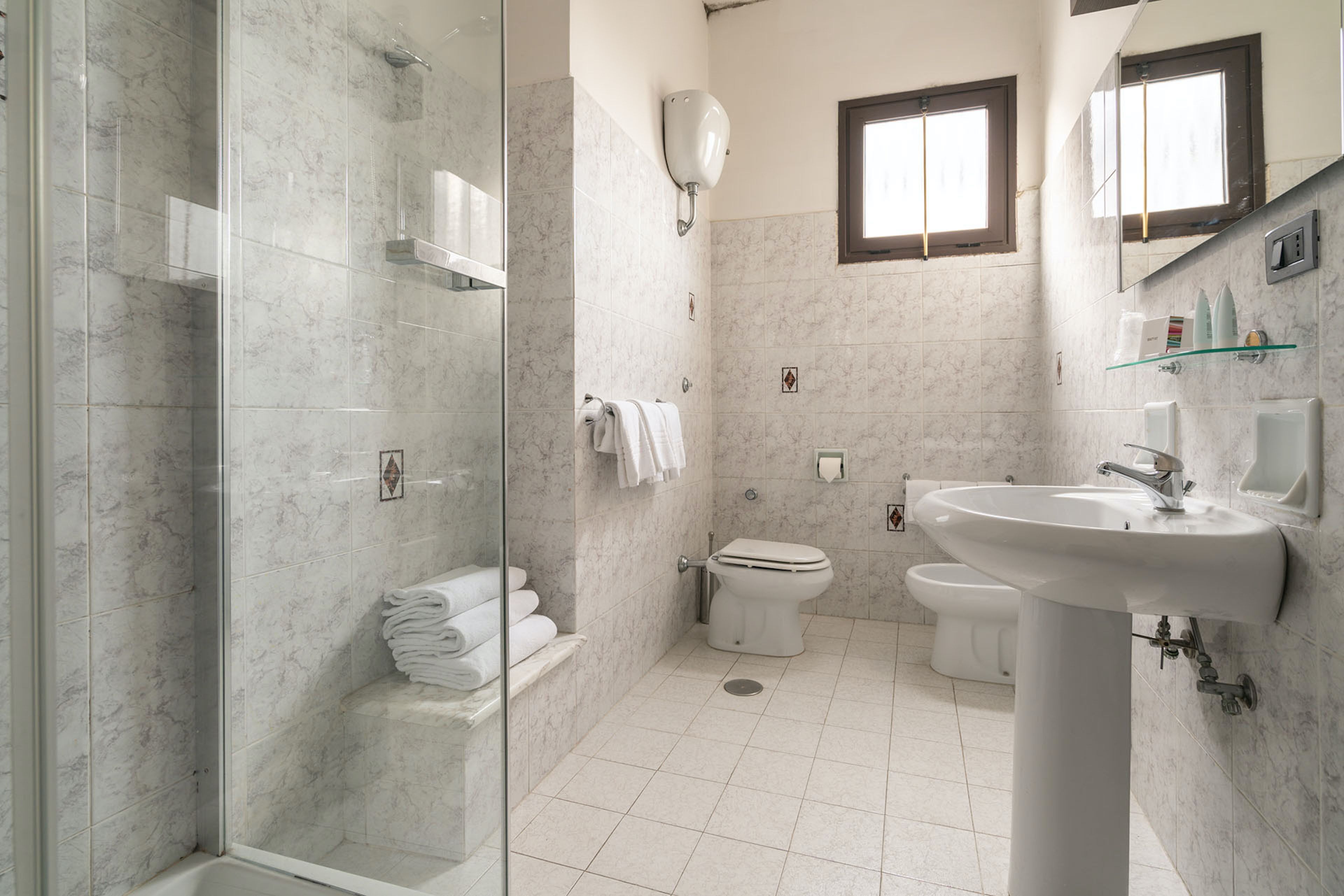 Een badkamer met een douche met glazen wand, toilet, wastafel en raam.