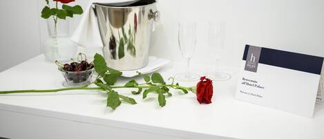 A white table with a silver pot, a glass vase with a red rose, a bowl of cherries, a wine glass, and a hotel welcome card.