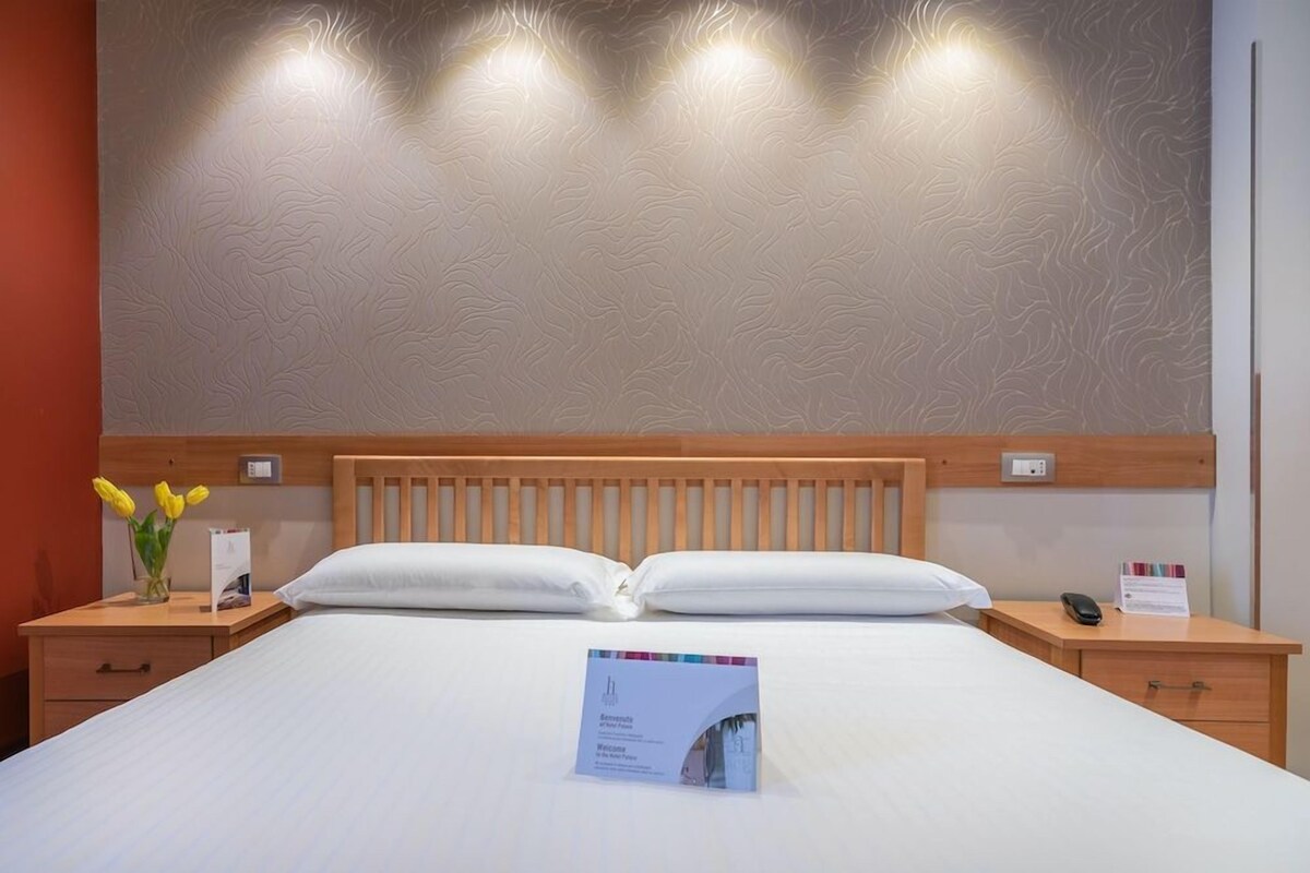 A hotel room with a wooden headboard, a bed with white linens, a bedside table with a vase of yellow flowers, a nightstand with a phone and a sign, and a textured wall with recessed lighting.