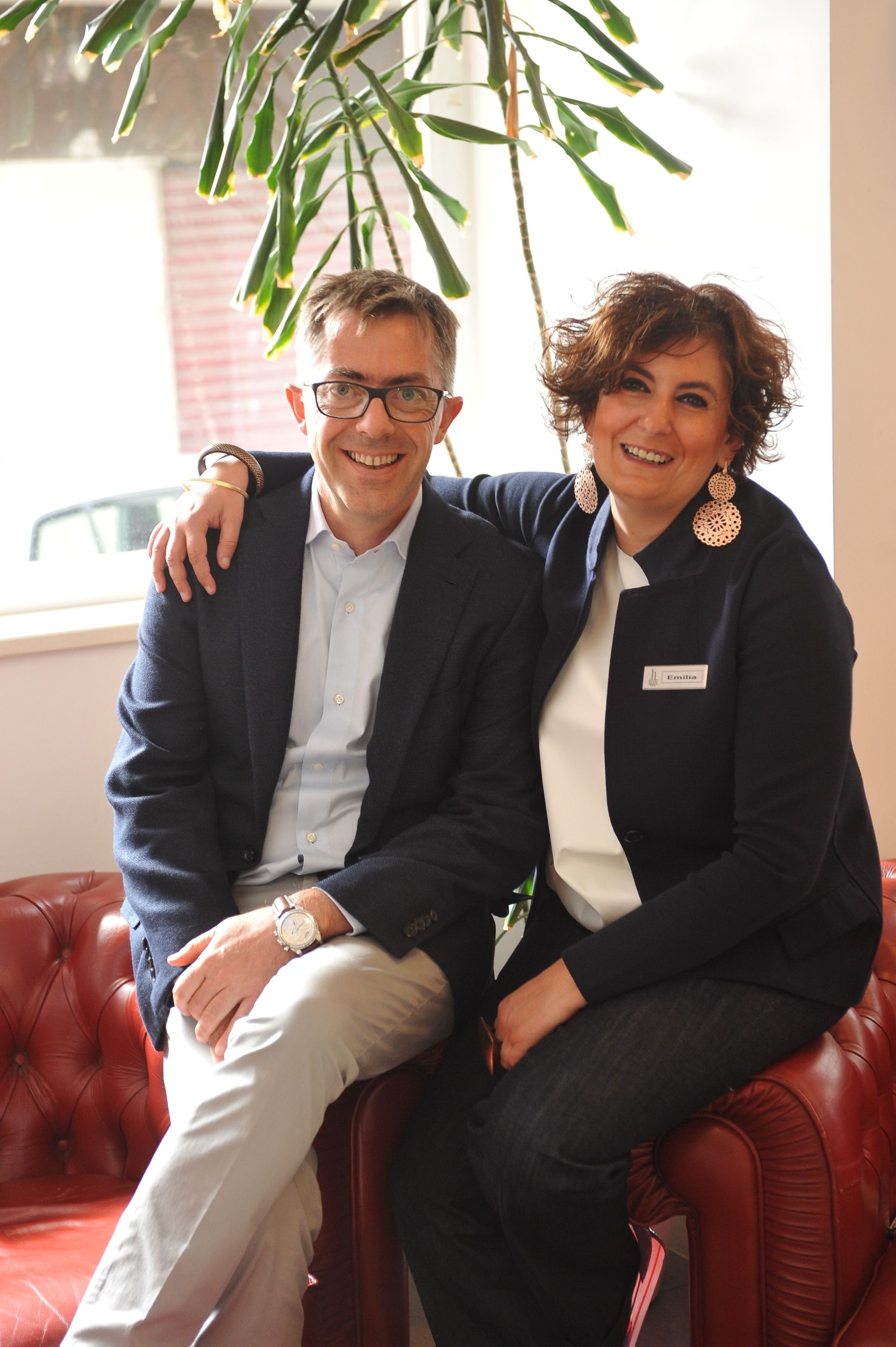 Two people sitting on a red leather couch in a room with a large window and a hanging plant.