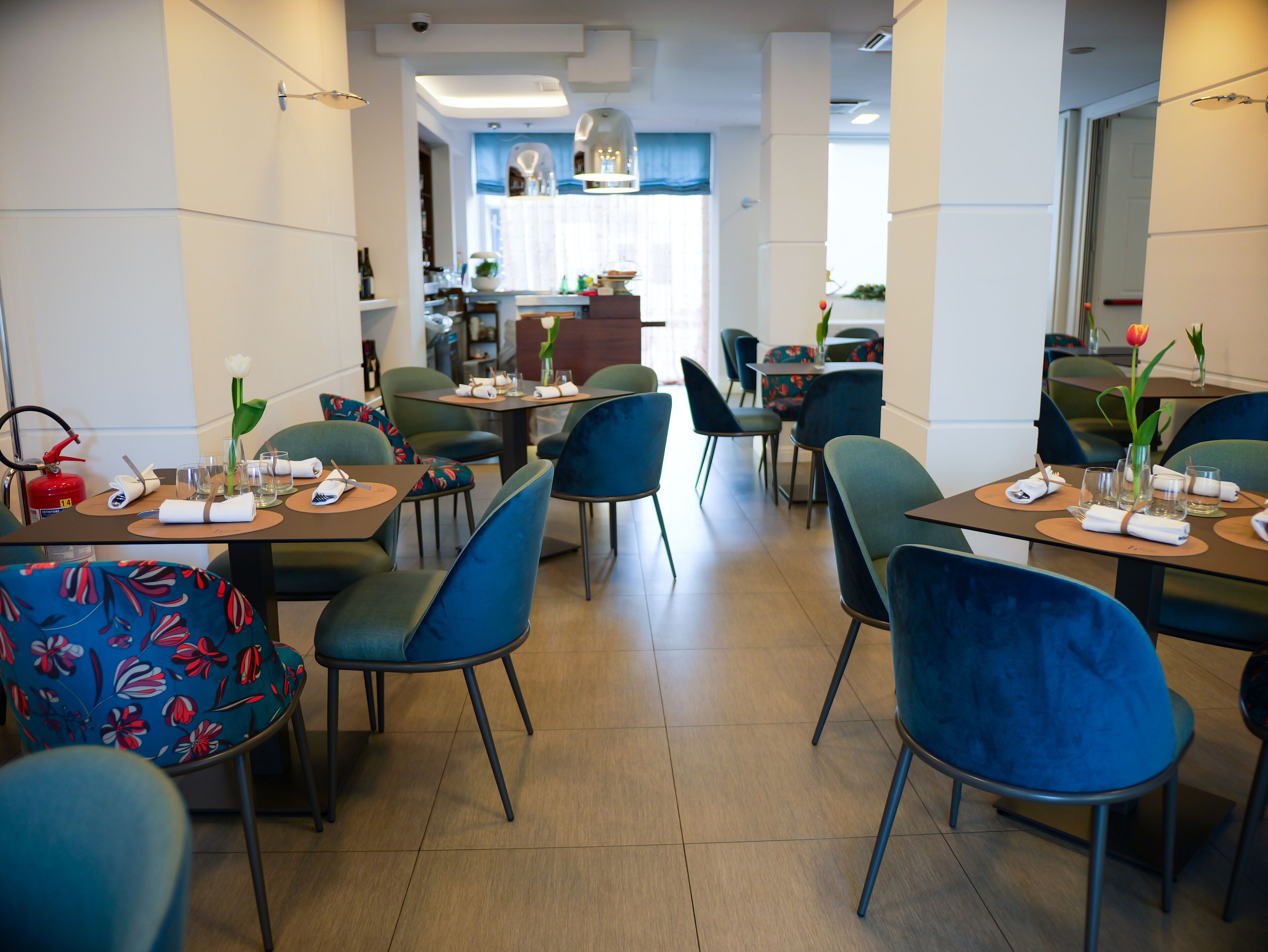 Un comedor moderno con varias mesas y sillas, una cocina visible al fondo y un extintor a la izquierda.