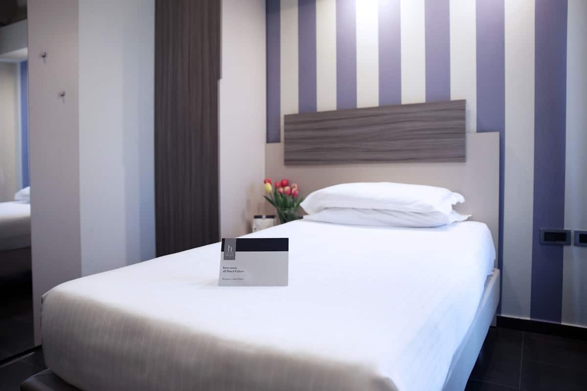 Ein ordentlich bezogenes Bett mit einem Schild, das Zimmerverfügbarkeit anzeigt, ein Nachttisch mit Blumenvase und ein gestreiftes Wanddesign.
