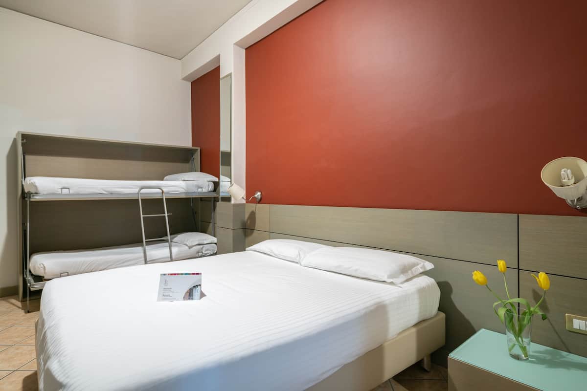 Ein Hotelzimmer mit Etagenbett, einem Einzelbett, einem Nachttisch mit Blumenvase und einer Wandlampe.