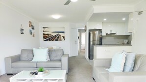 A modern living room with a grey sofa, a white coffee table, and a kitchen area with white cabinets and a stainless steel refrigerator. - Mari Court Resort (Surfers Paradise)