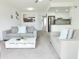 A modern living room with a grey sofa, a white coffee table, and a kitchen area with white cabinets and a stainless steel refrigerator.
