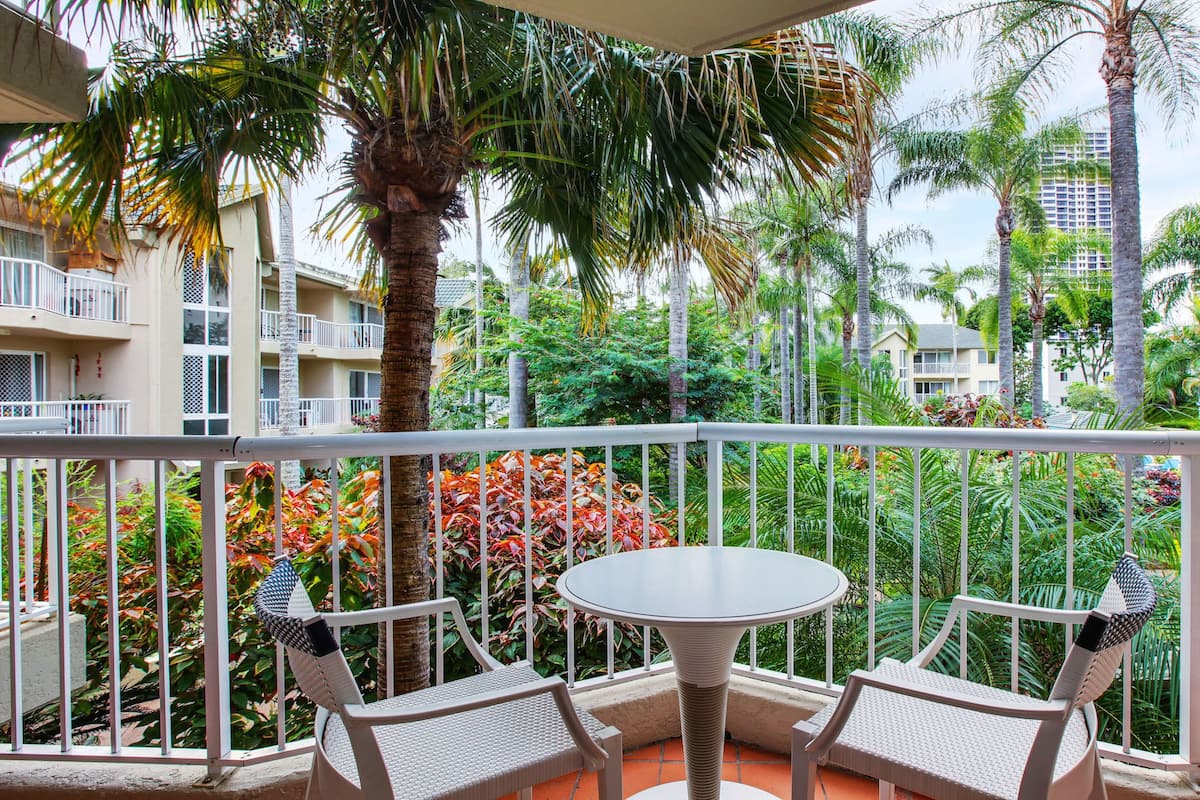 A balcony with two wicker chairs and a round table, surrounded by lush tropical plants and palm trees, overlooking a residential area with multi-story buildings.