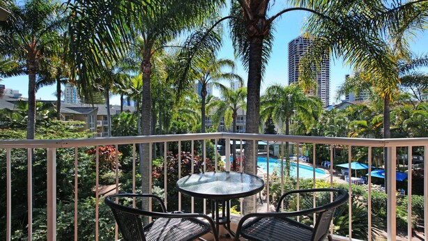 A balcony with a glass table and two chairs, overlooking a pool and palm trees. - Mari Court Resort (Surfers Paradise)