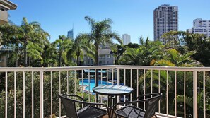 A balcony with a glass table and chairs, overlooking a swimming pool and palm trees, with tall buildings in the background. - Mari Court Resort (Surfers Paradise)