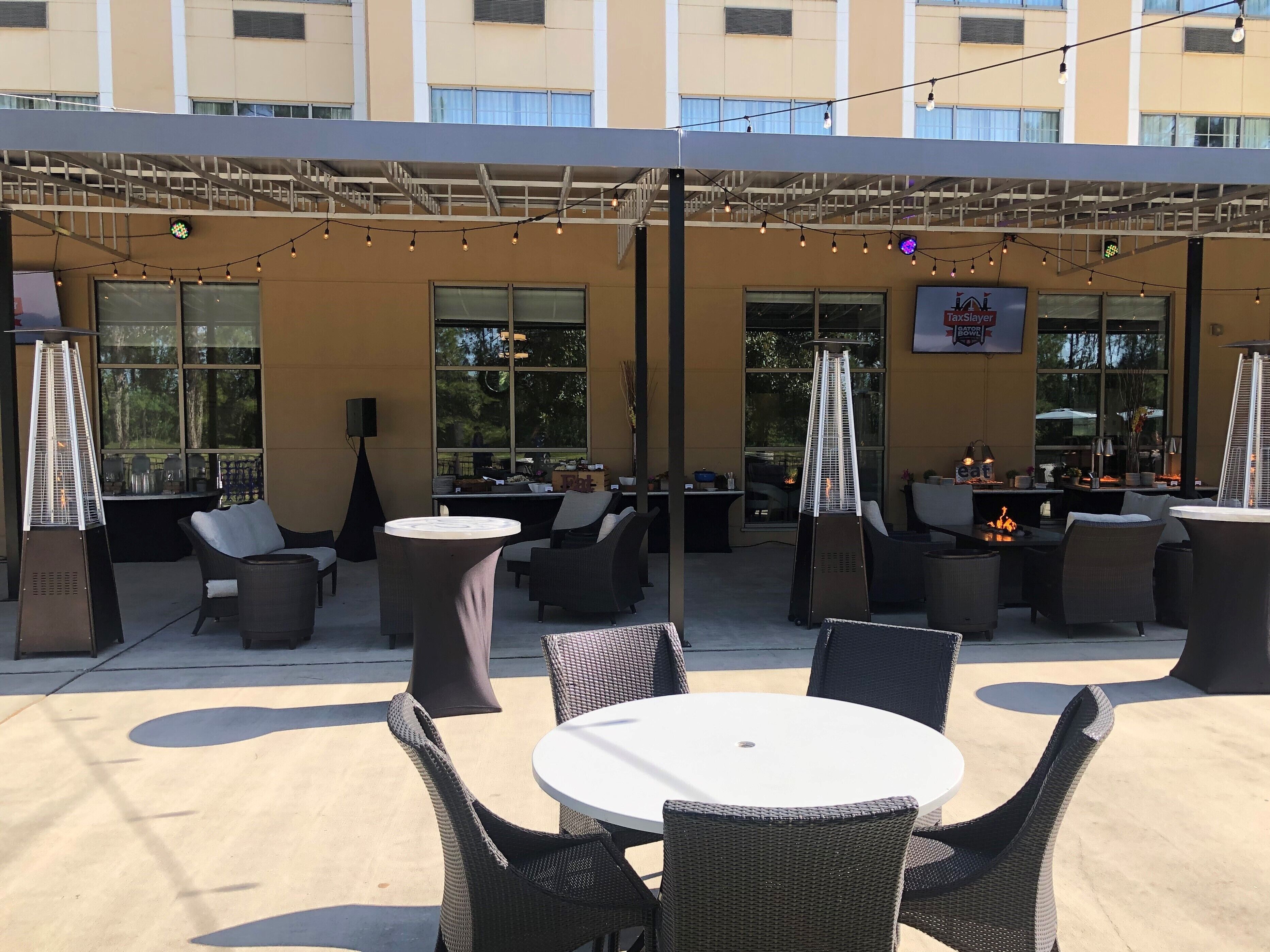 Un patio al aire libre con mesas redondas y sillas de mimbre, un escenario con luces colgantes y vistas a un edificio con grandes ventanales.