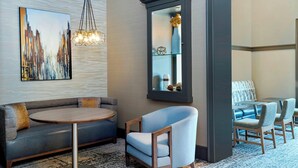 Un hall d’hôtel moderne avec un canapé gris, une table ronde en bois et un fauteuil bleu. Un grand tableau abstrait et un miroir avec un vase sont accrochés au mur.