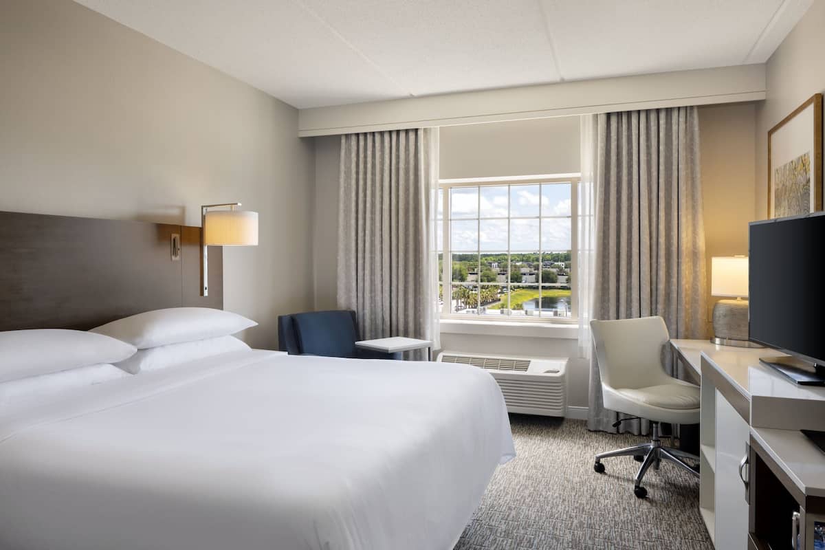 Habitación de hotel con una cama grande, un escritorio con computadora, una silla, un televisor y una ventana con cortinas.