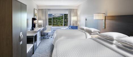 A hotel room with a large bed, a desk with a chair, a wardrobe, and a window with a view of greenery.