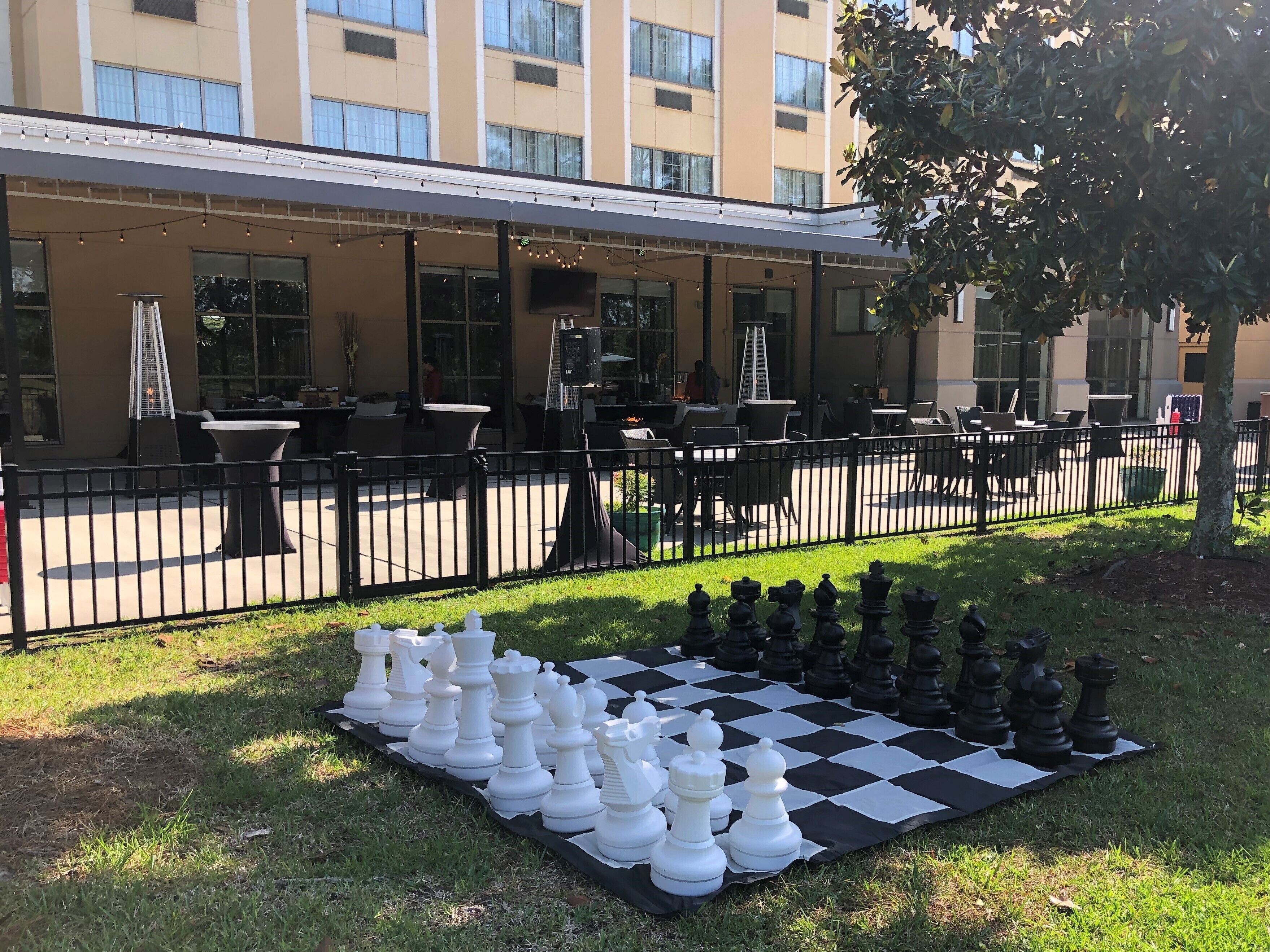 A large outdoor chess set on a checkered mat in front of a building with outdoor seating.