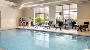 Une piscine intérieure à l’eau claire, entourée de chaises longues et de tables, avec de grandes fenêtres donnant sur la verdure environnante.