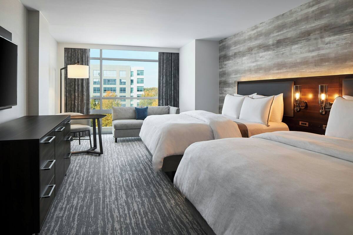 Ein Hotelzimmer mit zwei Betten, einem Schreibtisch, einem Sessel, einem Fernseher und einem großen Fenster mit Vorhängen.