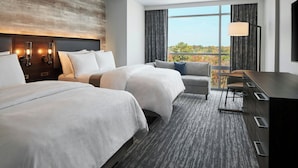 A hotel room with two beds, a sofa, a desk, and a large window offering a view of the outdoors.