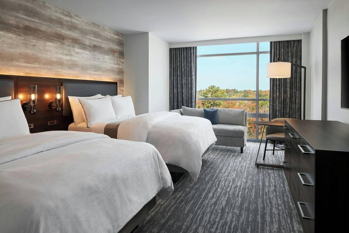 Ein Hotelzimmer mit zwei Betten, einem Sofa, einem Schreibtisch und einem großen Fenster mit Blick ins Freie.