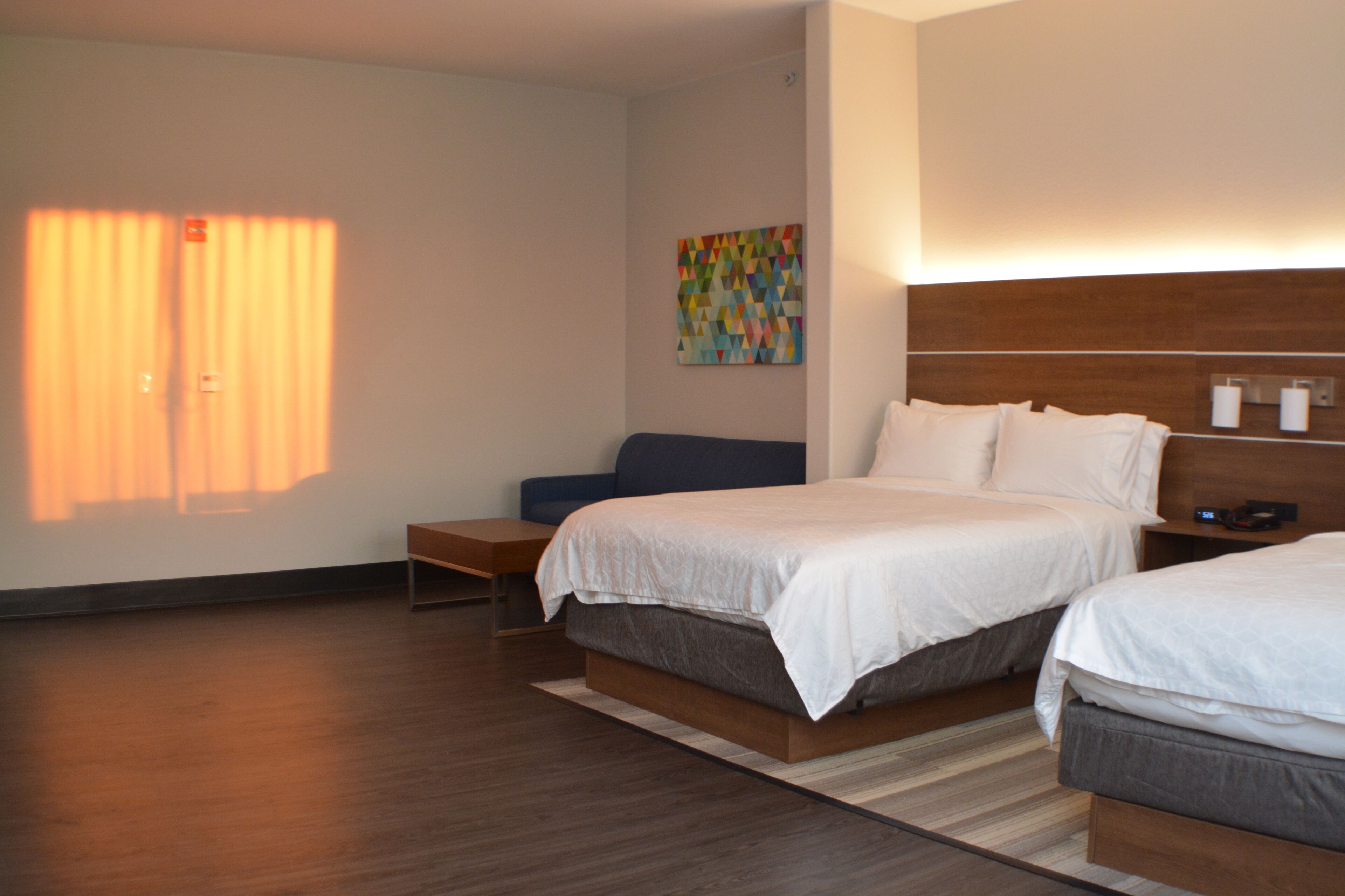 A modern hotel room with two beds, a wooden headboard, a colorful artwork, and a bench.