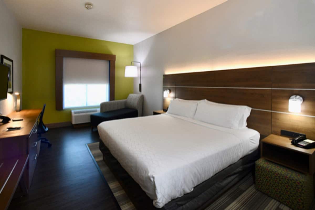 Une chambre d’hôtel avec un grand lit, un bureau avec un ordinateur, une chaise, une table de chevet avec une lampe et une fenêtre avec des stores.