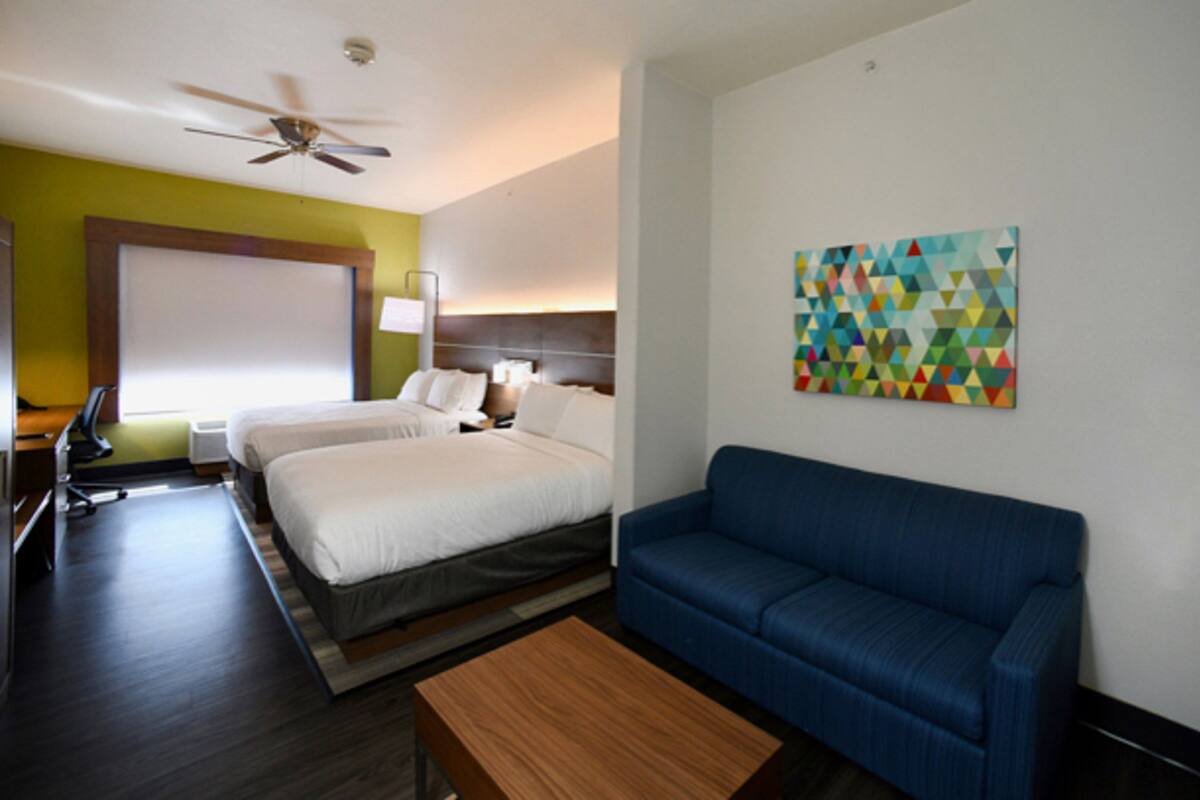 Une chambre d’hôtel avec un lit, un canapé bleu, un ventilateur de plafond, un bureau avec une lampe et une œuvre d’art murale aux motifs géométriques et aux couleurs vives.