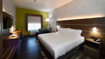 Quarto de hotel com uma cama grande, uma escrivaninha com computador, uma cadeira, uma mesa de cabeceira com abajur e uma janela com persianas.