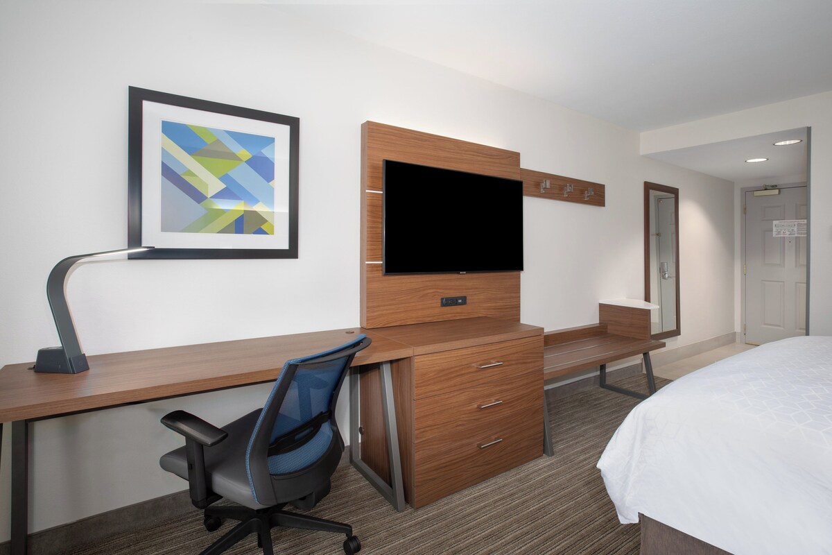 A hotel room with a wooden desk, a chair, a flat-screen TV mounted on a wooden cabinet, a bed with a white bedspread, and a framed abstract artwork on the wall.