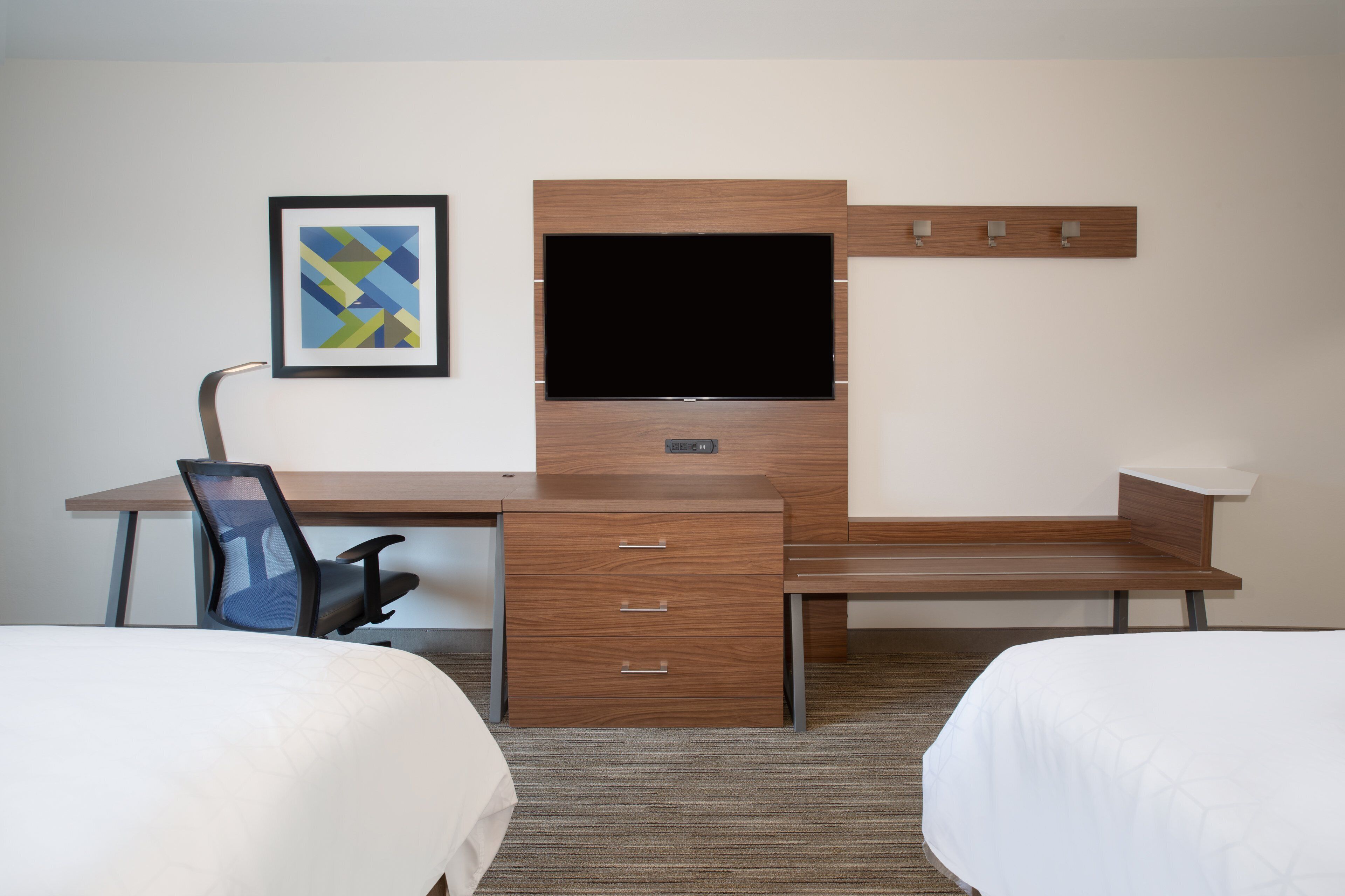 A hotel room with a wooden desk, a flat-screen TV, a chair, a bed, and a framed picture on the wall.