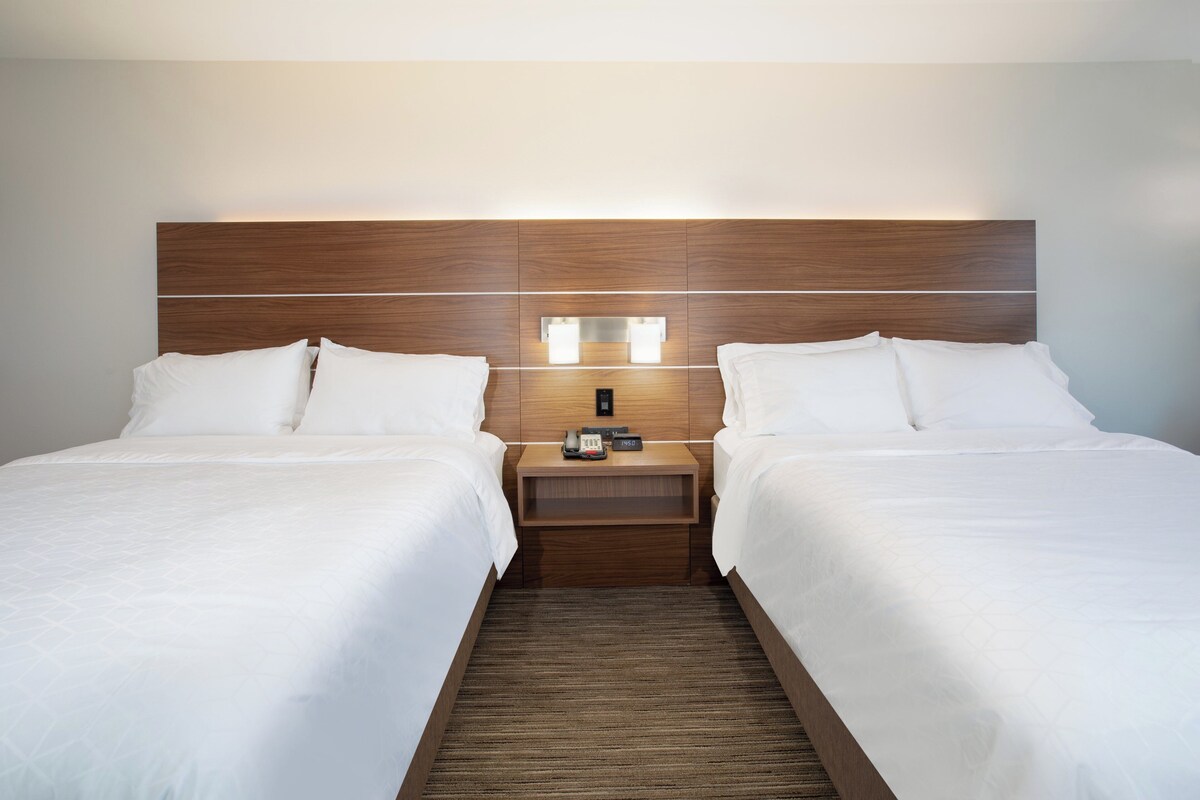 A hotel room with two beds, a wooden headboard, bedside lamps, and a nightstand with a phone and remote control.