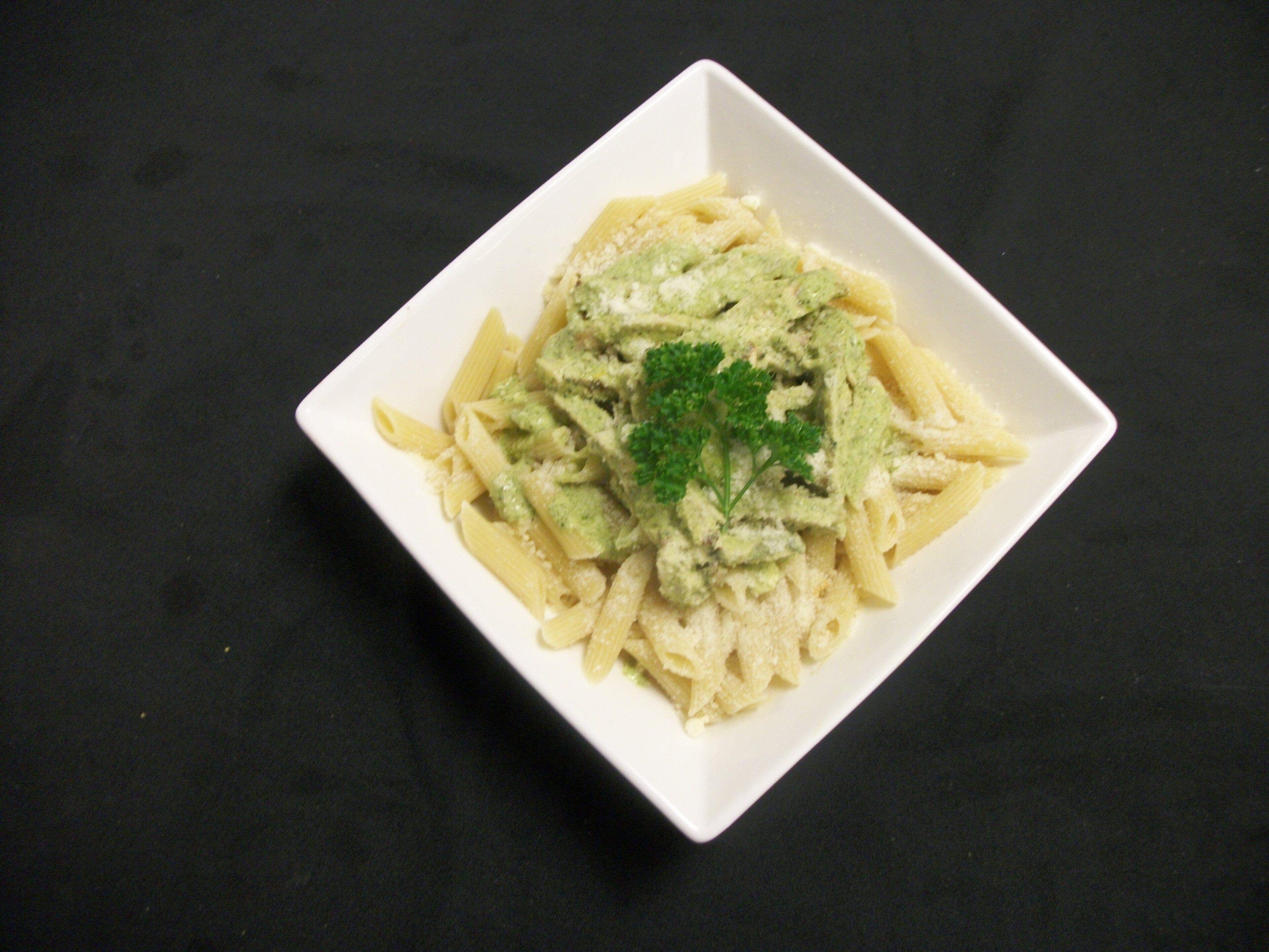 A square white plate with pasta and green sauce, garnished with parsley, on a dark surface.