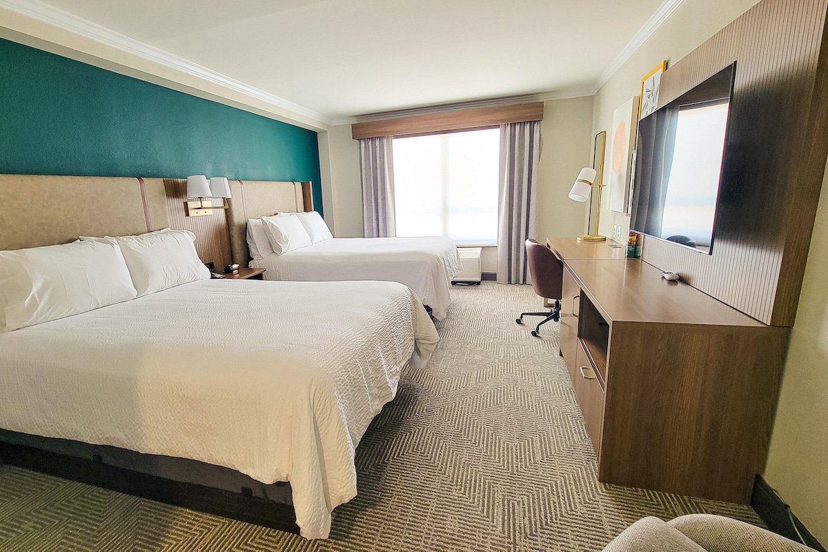 A hotel room with two beds, a desk, a chair, a TV, and a window with curtains.